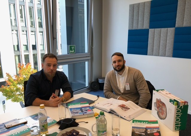 Deniz Tafrali und Sinan Barus am Besprechungstisch bei Elsevier in München, vor ihnen die MedAT-Bände und ein Anatomieatlas